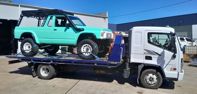 Teal lifted four-wheel drive with a roof rack strapped to the tilt tray outside a workshop