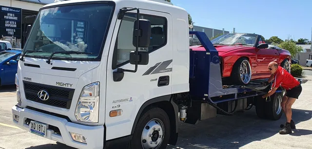 Driver tightening a wheel strap on a red convertible loaded on the tow truck