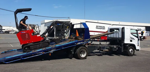 Red mini excavator being loaded onto the lowered tilt tray in a transport yard