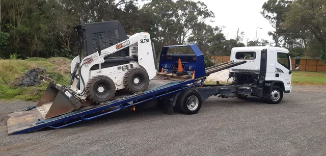 White skid steer loader with a bucket on the lowered tilt tray in a gravel yard